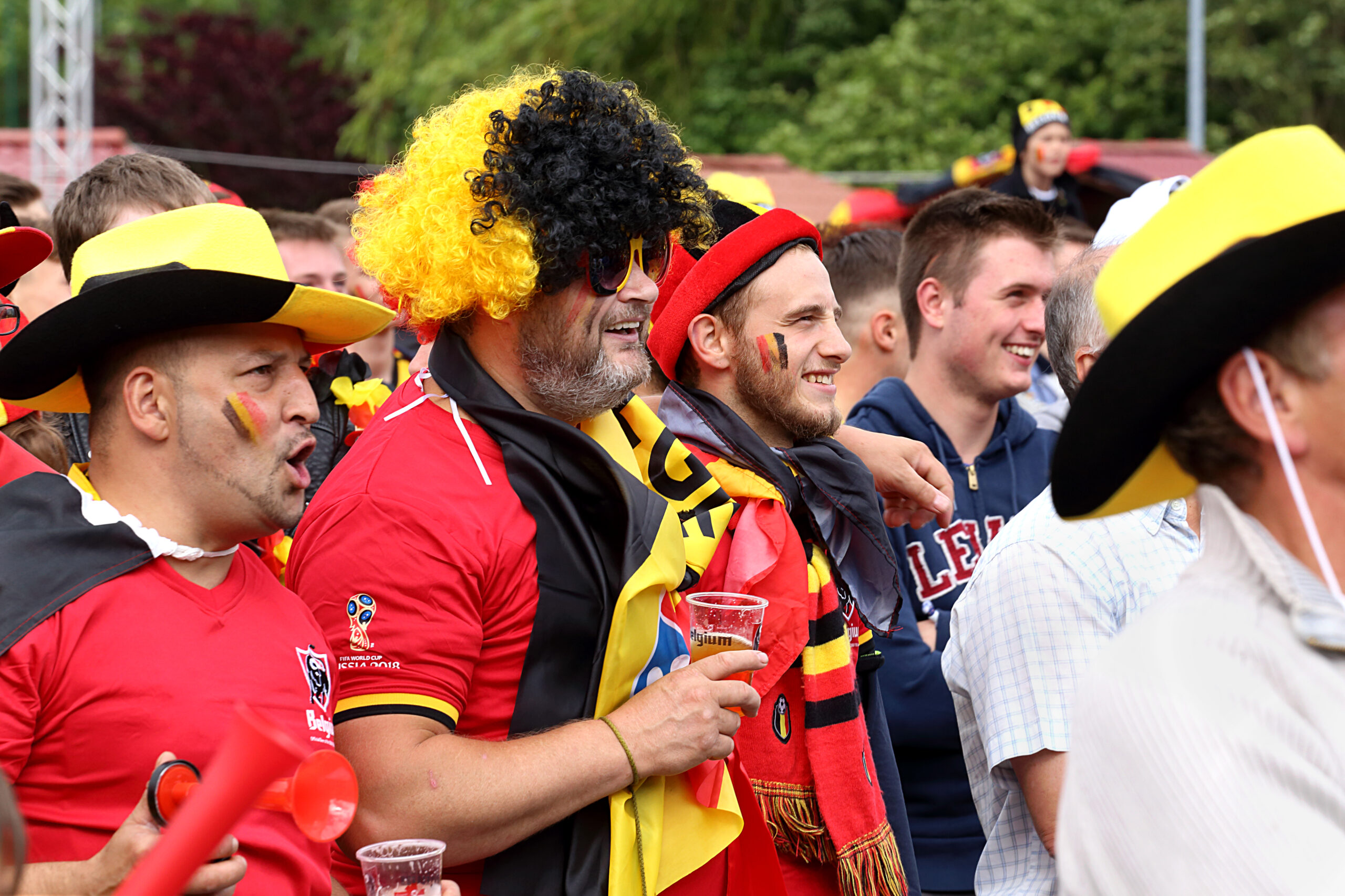 A crowd of soccer fans wearing team colors and flags, watching a match on a big outdoor screen